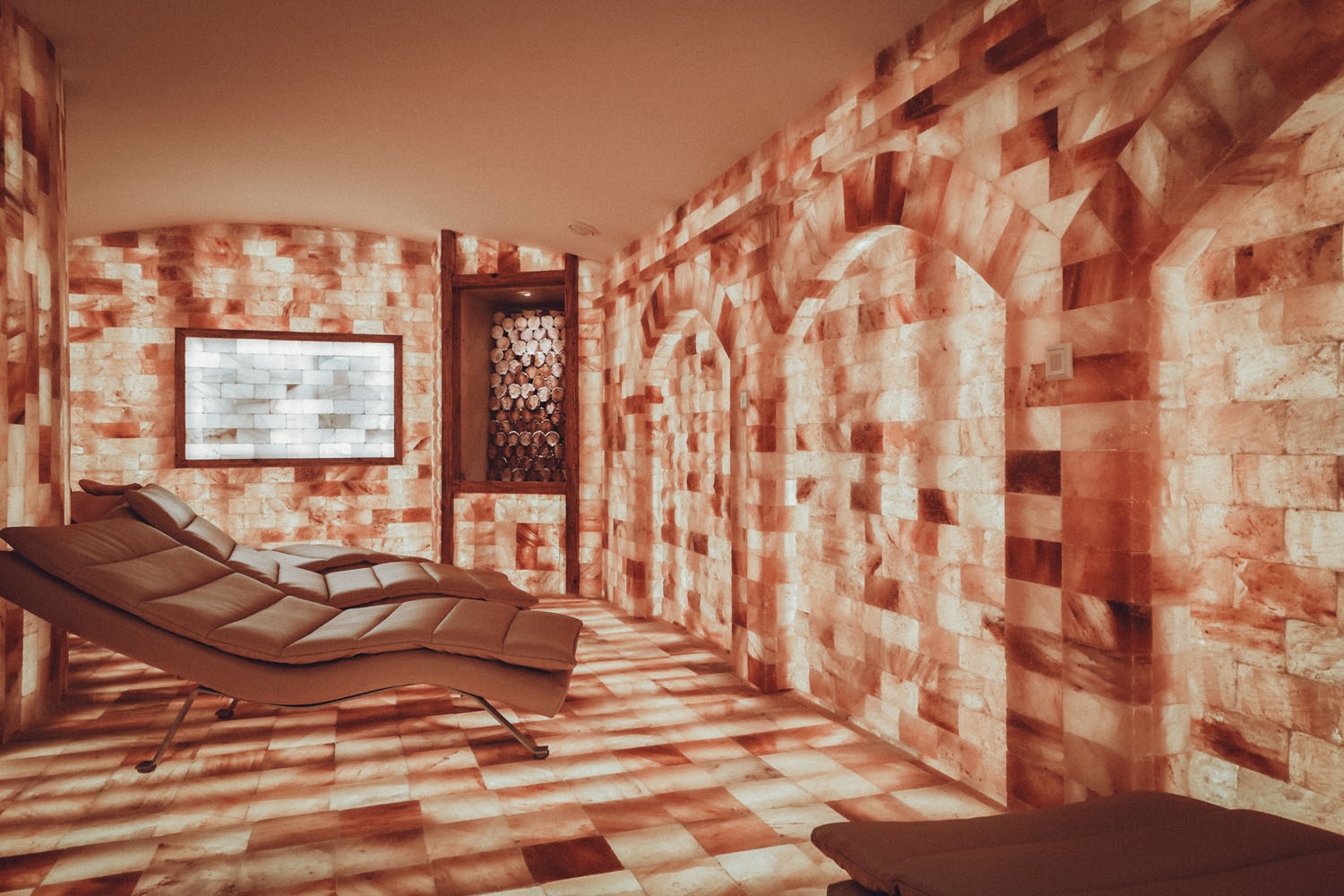 Ein Spa-Raum mit Liegestühlen und Wänden, Boden und Decke aus rosa Himalaya-Salzziegeln, die eine warme, leuchtende Atmosphäre schaffen. Ein Fenster lässt natürliches Licht herein.