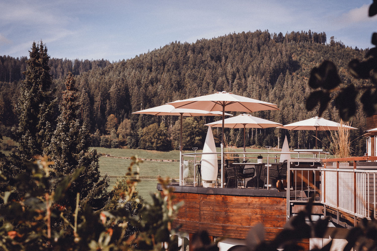 Eine Holzterrasse mit Tischen und Stühlen unter großen weißen Sonnenschirmen bietet an sonnigen Tagen einen Blick auf grüne Felder und bewaldete Hügel.