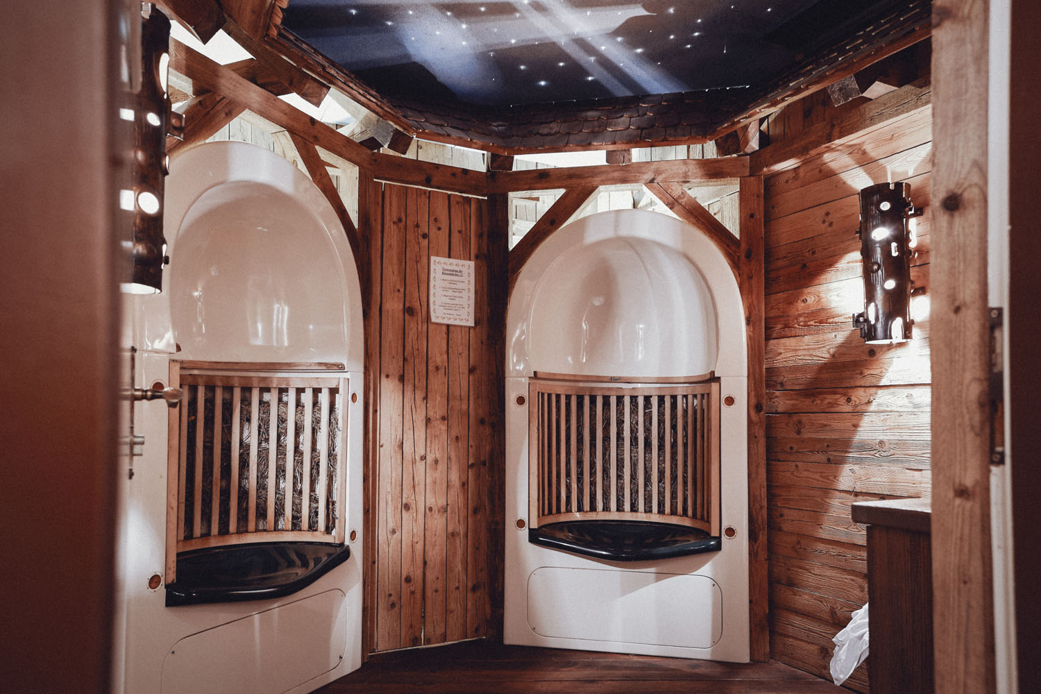 Ein gemütlicher Holzraum mit zwei weißen, kuppelförmigen Sitzecken, schwarzen Kissen und einem Sternenhimmel an der Decke. Die warme Beleuchtung und die rustikalen Holzpaneele schaffen eine entspannende, spaähnliche Atmosphäre.