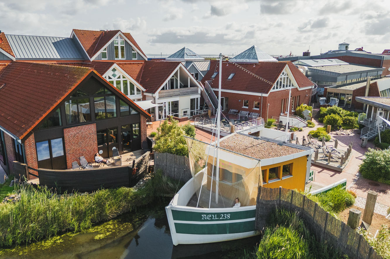 Luftaufnahme des Badewerks Neuharlingersiel. Ein reizvoller Gebäudekomplexes mit roten Dächern und großen Fenstern. Ein weiß-grünes Boot mit der Aufschrift „TÖL 238“ liegt an einem schmalen Wasserweg neben den Gebäuden vor Anker, umgeben von Grün an einem sonnigen Tag.