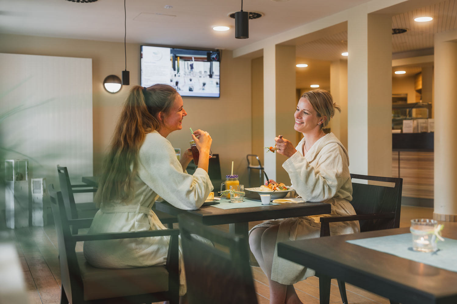 Zwei Frauen in weißen Gewändern sitzen an einem Tisch in einem modernen Restaurant, lächeln und genießen gemeinsam das Essen und die Getränke. Die Atmosphäre ist hell und entspannt, im Hintergrund sind Kerzen und ein Fernsehbildschirm zu sehen.