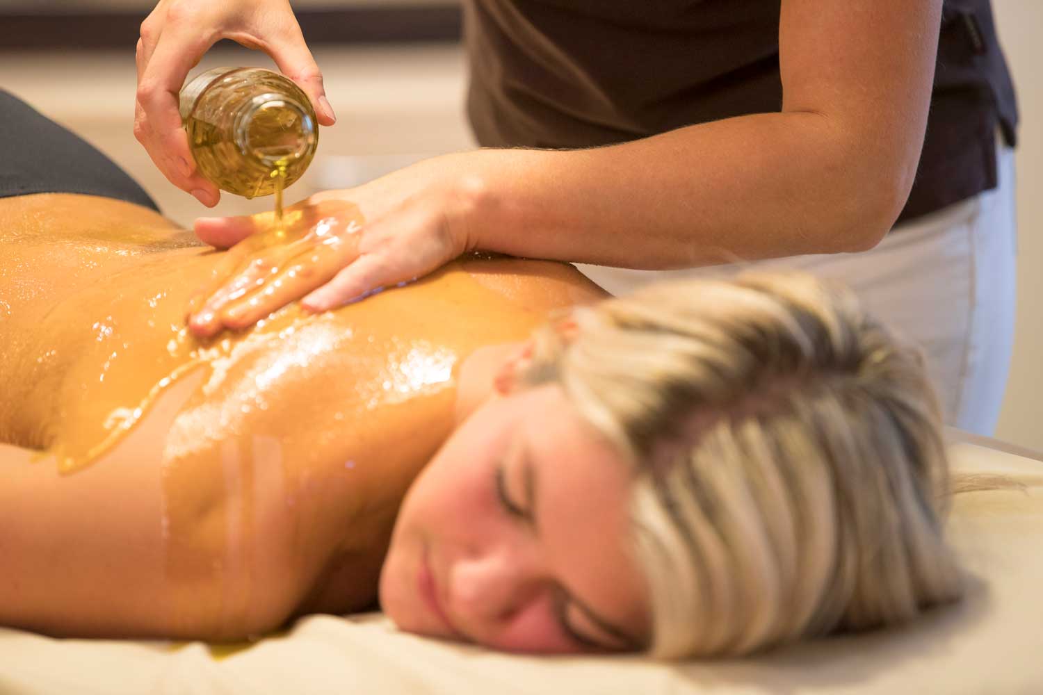 Eine Person liegt mit dem Gesicht nach unten auf einer Massageliege, während eine andere Person Öl auf ihren Rücken gießt und ihn massiert, wodurch eine entspannende Spa-Atmosphäre entsteht.