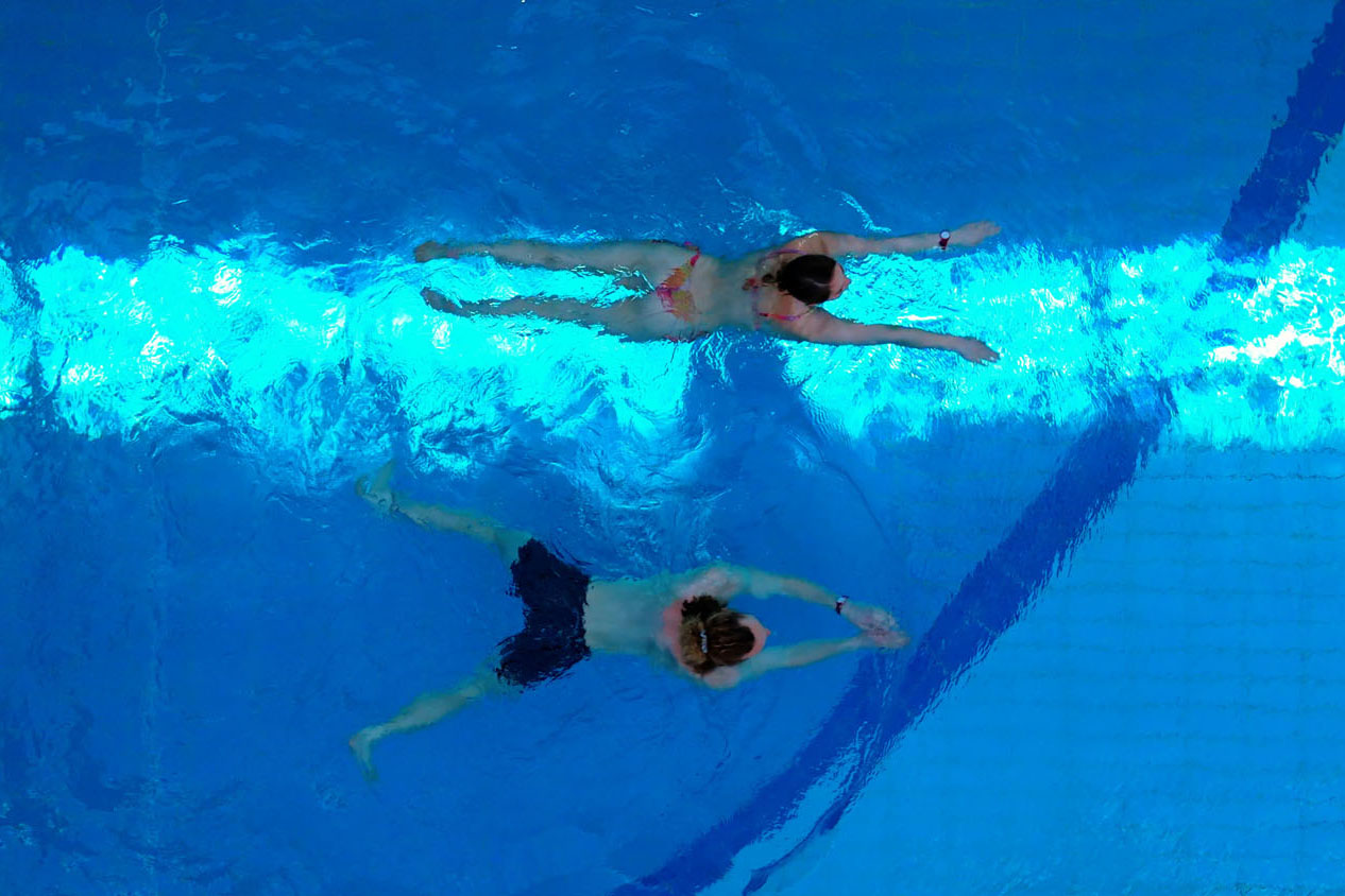 Zwei Personen schwimmen nebeneinander in einem Schwimmbecken, von oben gesehen. Die eine trägt einen rosa Badeanzug, die andere eine blaue Badehose. Beide strecken ihre Arme nach vorne und schwimmen entlang einer hellen Bahnmarkierung.