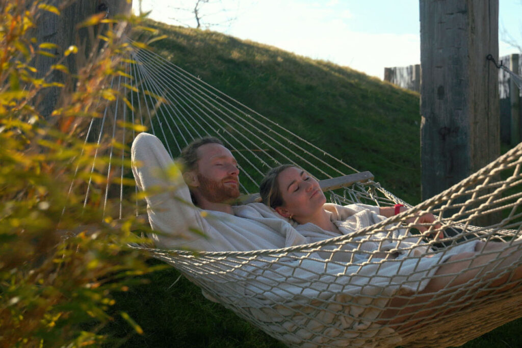 Ein Mann und eine Frau entspannen sich gemeinsam im Freien in einer Netzhängematte; beide tragen leichte Gewänder. Sie wirken ruhig und zufrieden und genießen die Sonne und das Grün um sich herum.