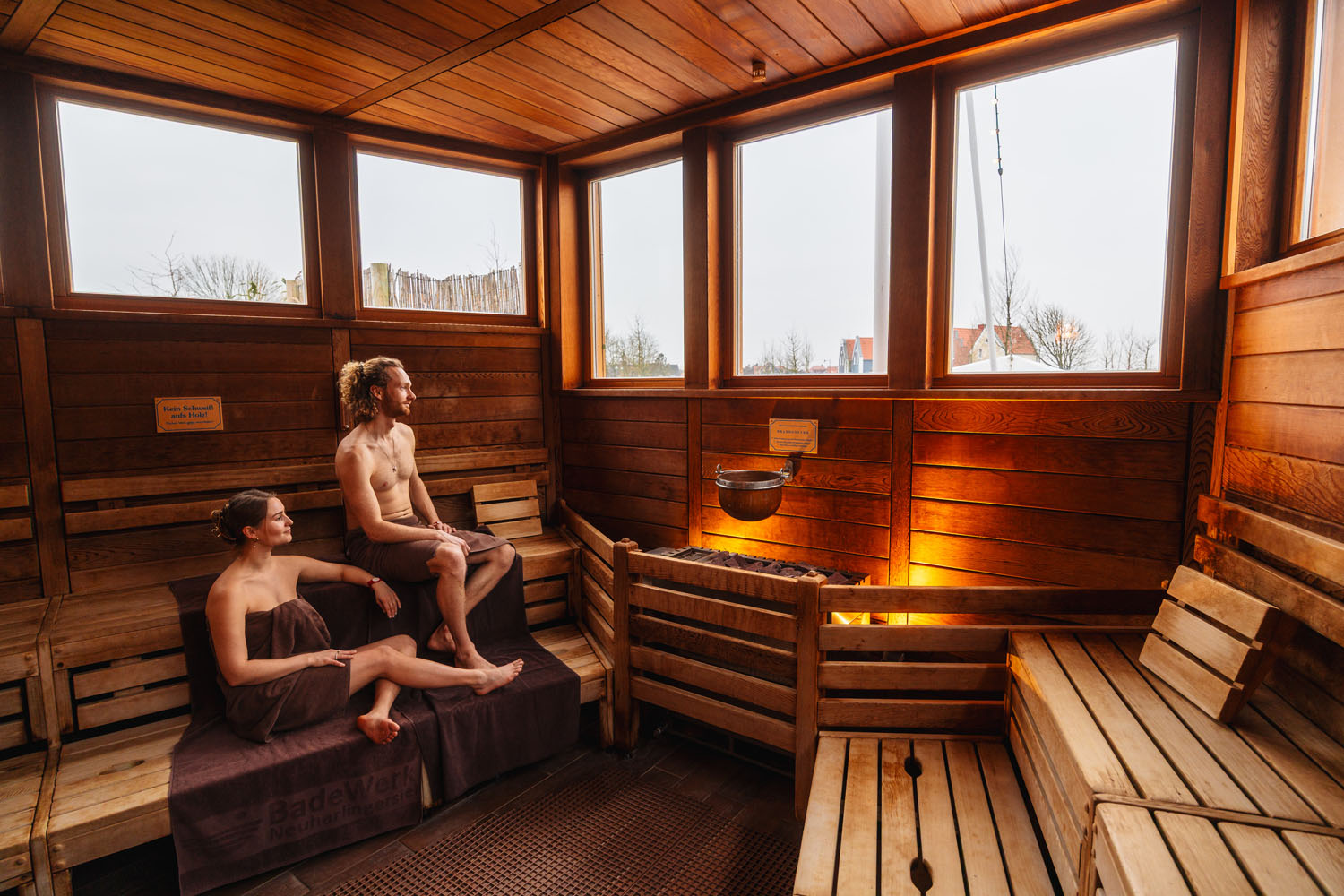 Zwei Personen sitzen in einer Sauna auf Holzbänken und sind in Handtücher gewickelt. Die Sauna besteht aus hellem Holz und verfügt über große Fenster, durch die Tageslicht hereinfällt. Über den erhitzten Steinen in der Mitte hängt ein Metalleimer.