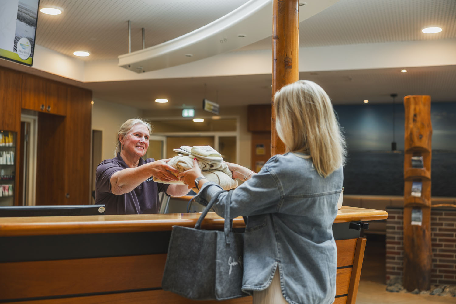 Eine lächelnde Rezeptionistin reicht einer Frau mit blonden Haaren an der Rezeption Handtücher. Die Frau hält eine graue Tasche in der Hand und trägt eine Jeansjacke. Die Umgebung wirkt mit ihrer Holzausstattung und der sanften Beleuchtung warm und einladend.
