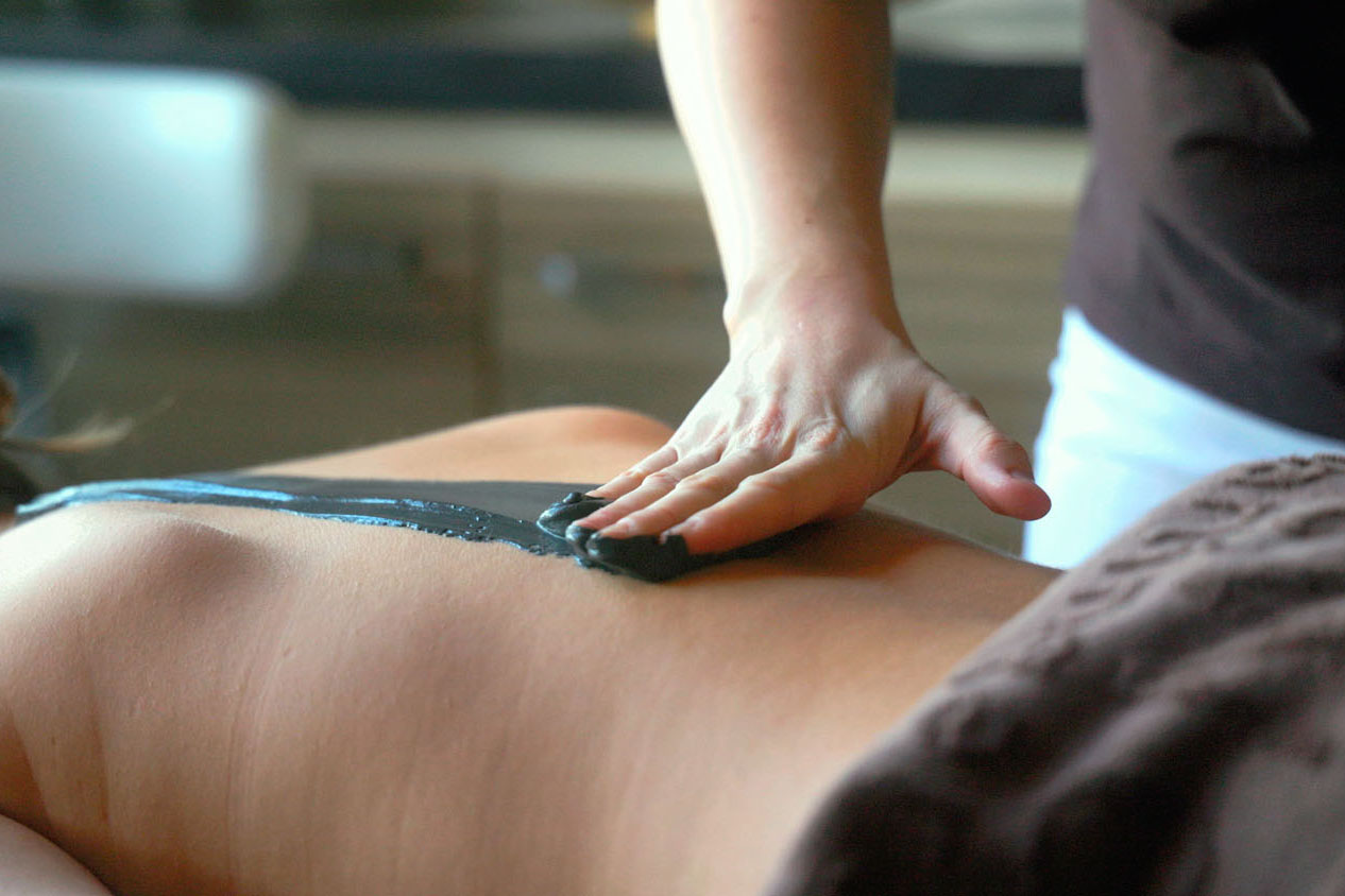 Eine Person trägt während einer Spa-Behandlung eine dunkle Schlamm- oder Tonmaske auf den nackten Rücken einer anderen Person auf, wobei ein Handtuch einen Teil des Körpers bedeckt. Der Hintergrund ist unscharf.