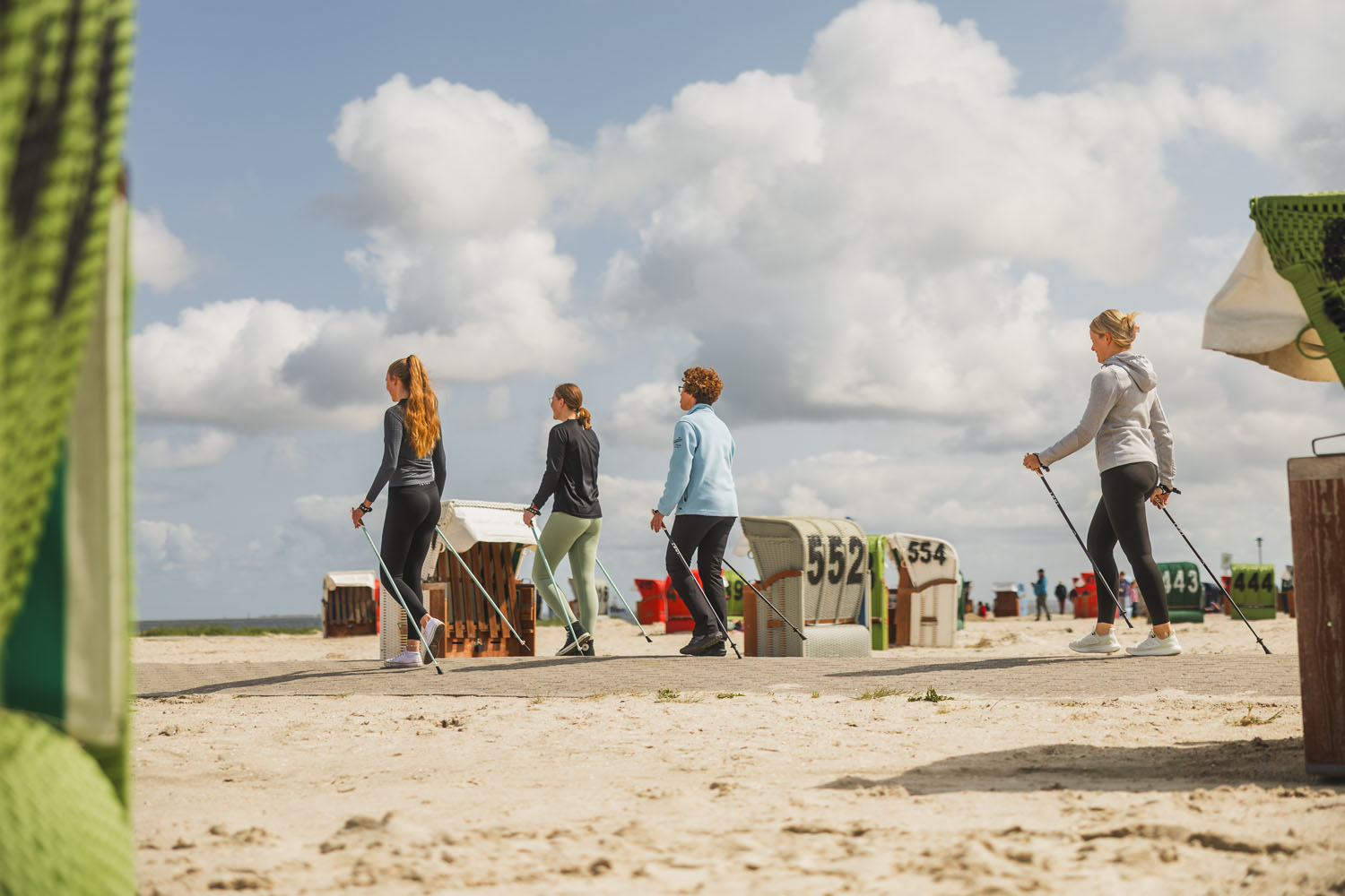 Vier Frauen gehen mit Stöcken am Sandstrand Nordic Walking und kommen unter einem teilweise bewölkten Himmel an gestreiften Strandliegen vorbei.