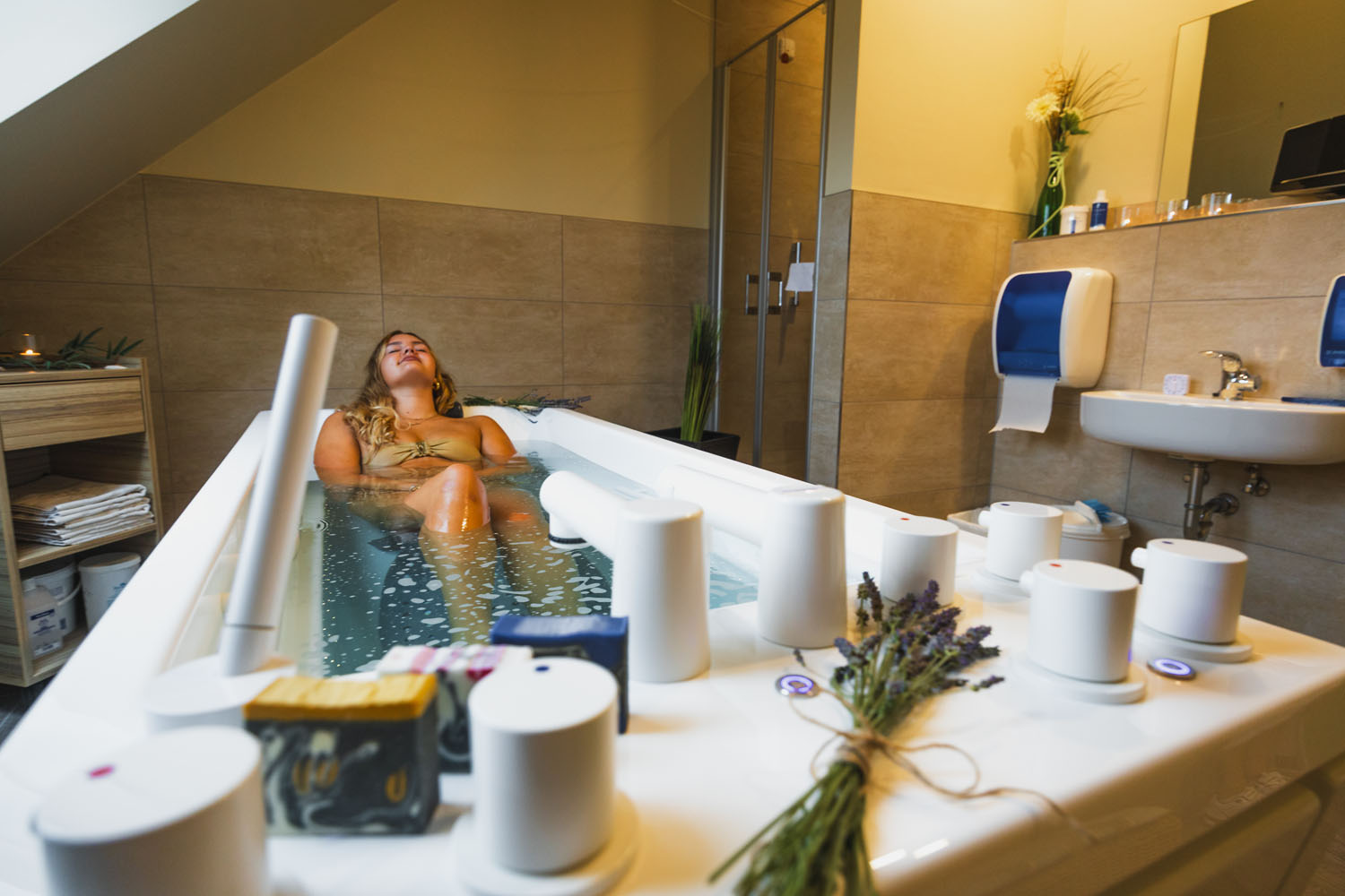 Eine Frau entspannt sich in einer modernen, mit Wasser gefüllten Badewanne in einem Badezimmer im Spa-Stil. Auf dem Wannenrand liegen Lavendel, Seife und Spa-Produkte, und der Raum besticht durch eine warme Beleuchtung und beige geflieste Wände.