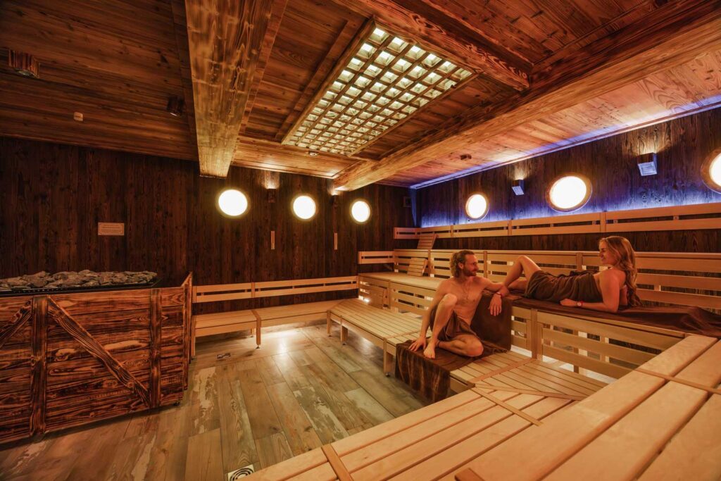 Zwei Menschen entspannen sich auf Holzbänken in einer gemütlichen, schwach beleuchteten Sauna mit warmer Beleuchtung, Holzwänden und -decke sowie einem mit Steinen gefüllten Ofen in der Ecke.