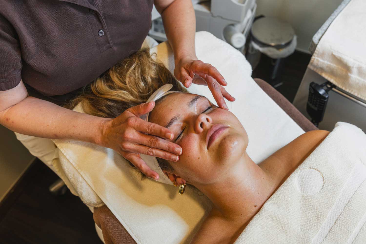 Eine Frau liegt mit geschlossenen Augen auf einer Spa-Liege und lässt sich von einer Kosmetikerin das Gesicht massieren. Die Hände der Kosmetikerin üben sanften Druck auf die Stirn und die Schläfen der Frau aus. Im Hintergrund sind Spa-Geräte zu sehen.