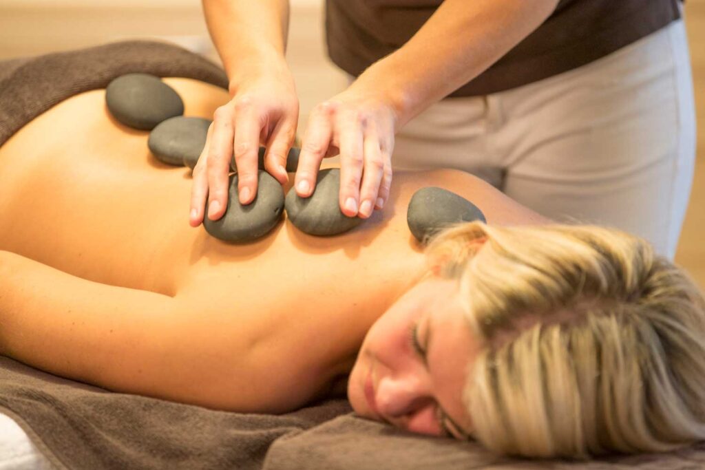 Eine Person liegt mit geschlossenen Augen mit dem Gesicht nach unten auf einer Massageliege, während ein Masseur ihr im Rahmen einer Hot-Stone-Massage warme Steine auf den Rücken legt.