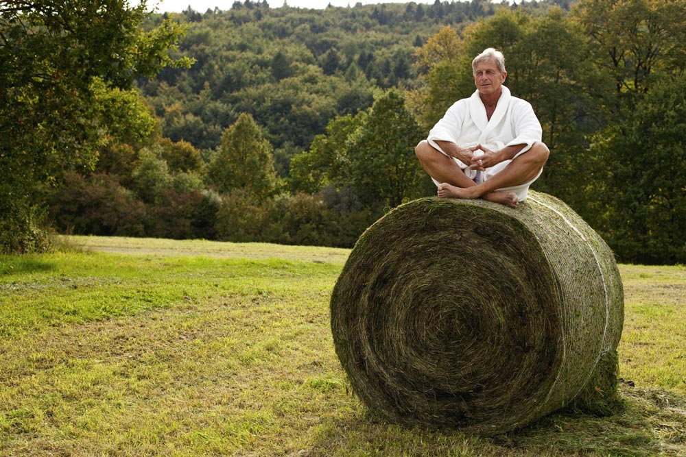 Ein älterer Mann in einem weißen Gewand sitzt mit gekreuzten Beinen auf einem großen runden Heuballen auf einer Wiese und meditiert, im Hintergrund sind Bäume und Hügel zu sehen.