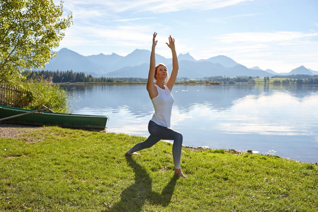 Eine Frau praktiziert Yoga im Freien in einer Ausfallschritt-Pose mit erhobenen Armen auf grünem Gras am Ufer eines ruhigen Sees, im Hintergrund sind Berge und ein strahlend blauer Himmel zu sehen.