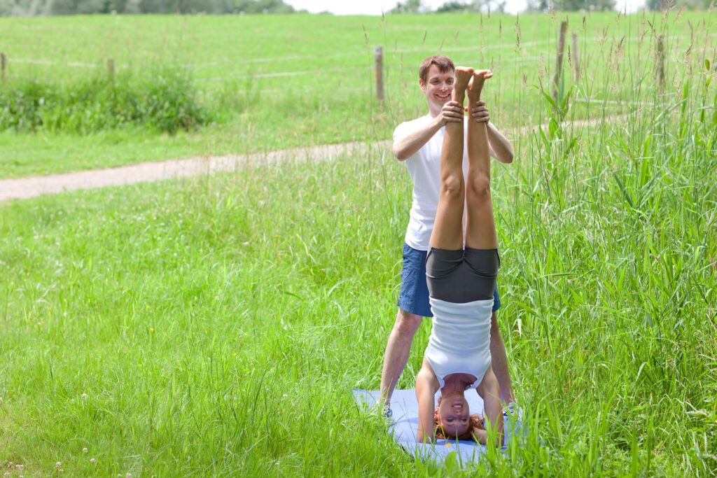 Ein Mann hilft einer Frau dabei, in einer Yoga-Kopfstand-Pose auf einer Matte im Freien auf einer Wiese das Gleichgewicht zu halten, im Hintergrund sind Grünflächen und ein Feldweg zu sehen.