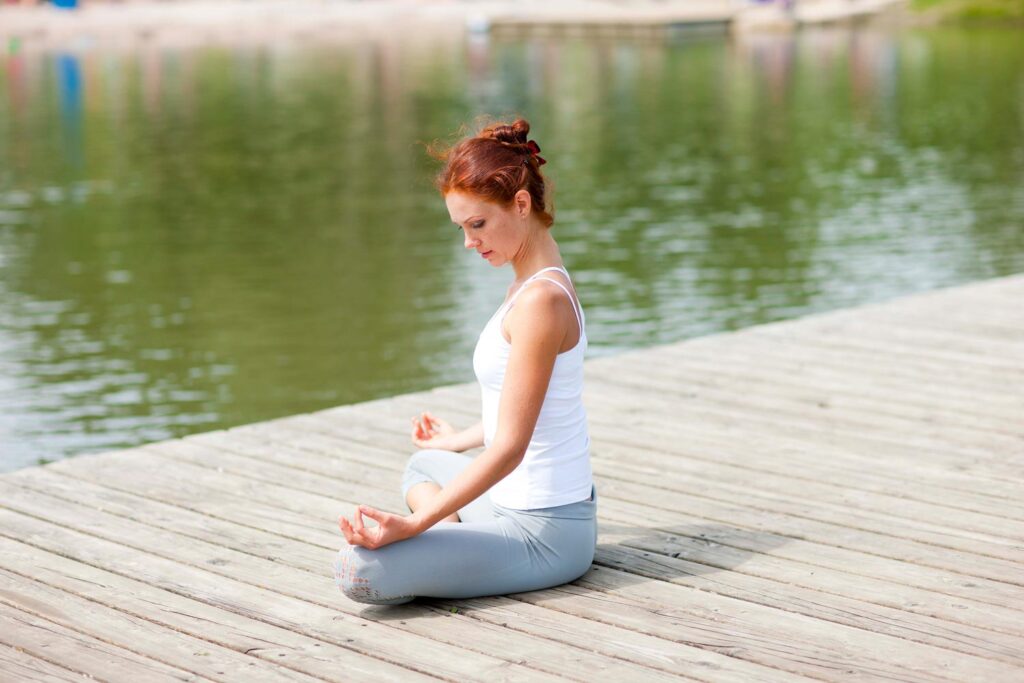 Eine rothaarige Frau, die ein weißes Tanktop und graue Leggings trägt, sitzt im Schneidersitz in einer meditativen Haltung auf einem Holzsteg an einem ruhigen Gewässer.