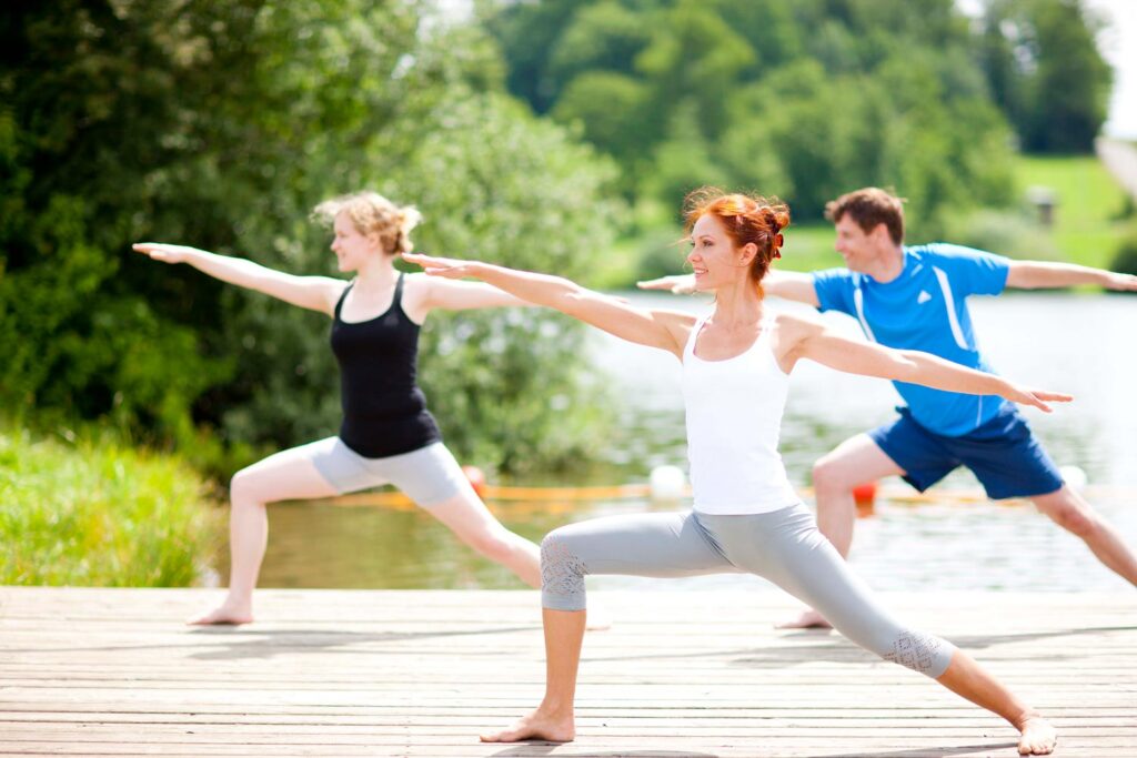 Drei Menschen praktizieren Yoga im Freien auf einer Holzterrasse in der Nähe eines Sees und nehmen dabei Krieger-Posen ein. Im Hintergrund sind an einem sonnigen Tag Bäume und Grünflächen zu sehen.
