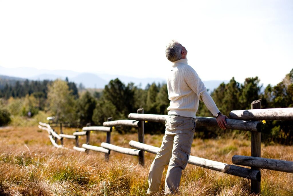 Ein älterer Mann in einem weißen Pullover lehnt sich auf einer Wiese an einen Holzzaun und blickt in den Himmel. Im Hintergrund sind unter einem strahlend blauen Himmel Bäume und Berge zu sehen.