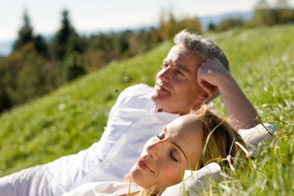 Ein Mann und eine Frau entspannen sich auf einem grasbewachsenen Hügel; sie liegen nebeneinander im Sonnenlicht, die Augen geschlossen, und wirken friedlich und zufrieden. Im Hintergrund sind Bäume und ein blauer Himmel zu sehen.