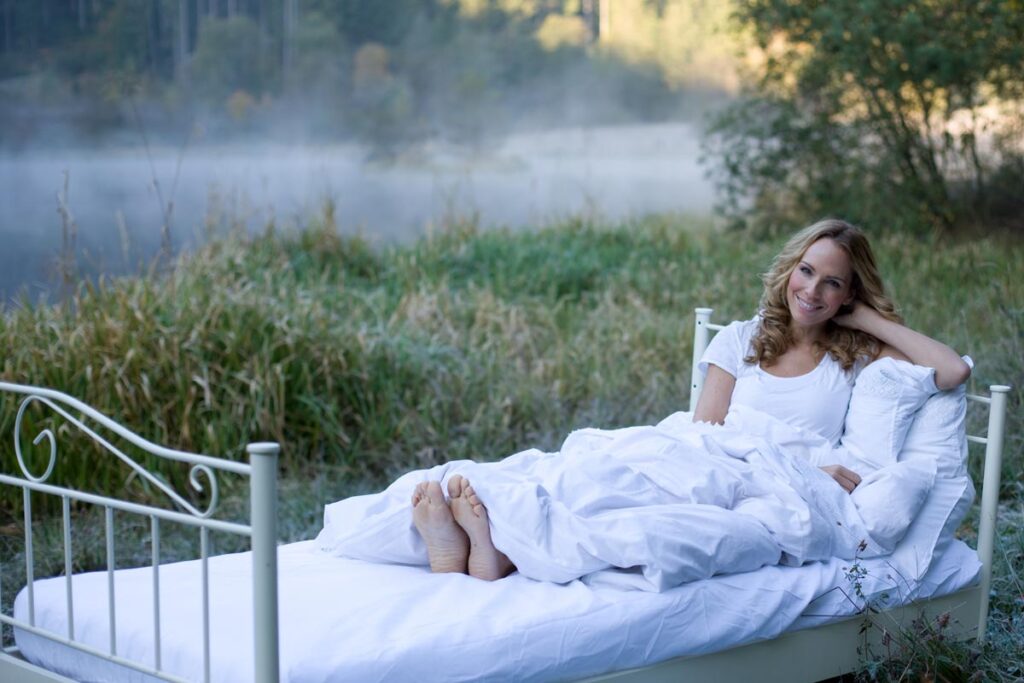 Eine Frau in einem weißen Pyjama lächelt, während sie sich im Freien im Bett aufrichtet und mit einer weißen Decke zugedeckt ist. Das Bett steht auf einer Wiese neben einem nebelverhangenen See und ist von Bäumen umgeben.