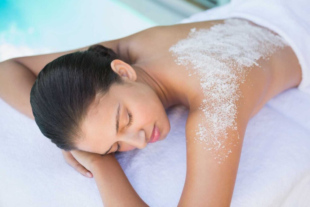 Eine Frau mit dunklem Haar liegt mit geschlossenen Augen auf dem Bauch auf einer Massageliege und entspannt sich. Auf ihrem nackten Rücken befindet sich eine Schicht aus Salz oder einem Peeling, was auf eine Wellness-Behandlung hindeutet.