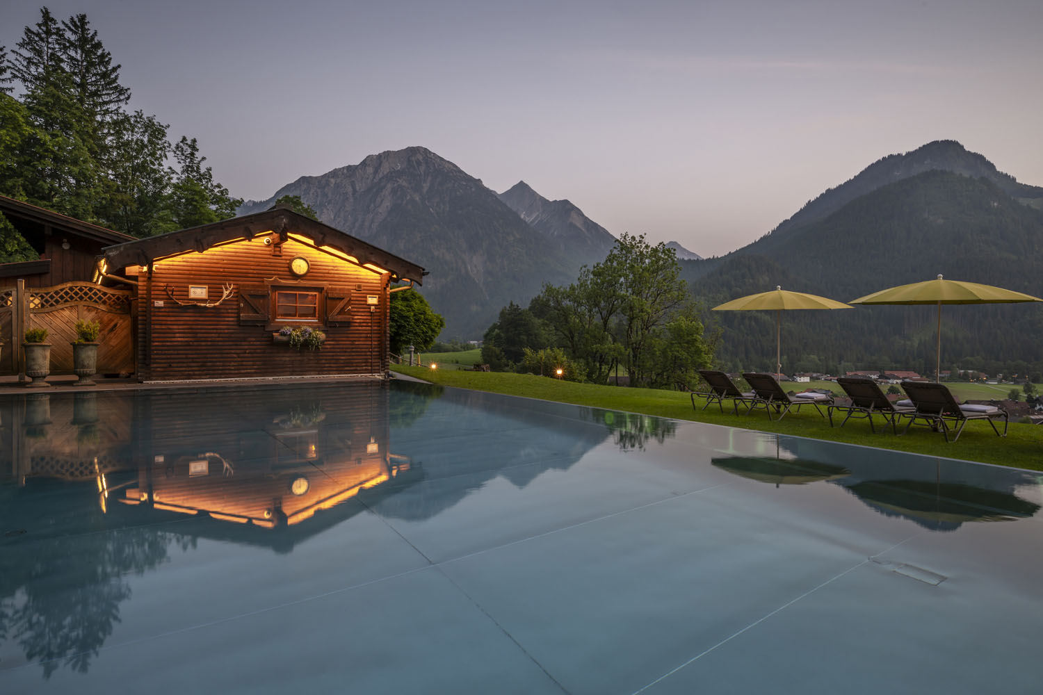 In einem ruhigen Außenpool spiegelt sich eine Holzhütte wider, im Hintergrund sind Berge und Bäume zu sehen. Bei Sonnenuntergang säumen mehrere Liegestühle und gelbe Sonnenschirme den Poolrand und schaffen so eine friedliche, malerische Kulisse.
