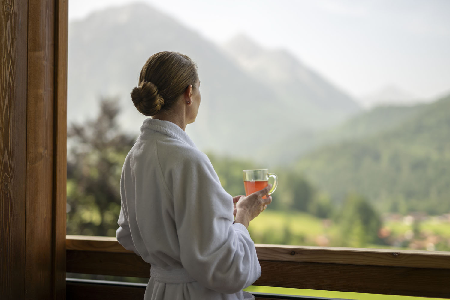 Eine Frau in einem weißen Bademantel steht auf einem Balkon, hält eine Tasse Tee in der Hand und blickt an einem dunstigen Tag auf eine malerische Landschaft aus grünen Hügeln und fernen Bergen.