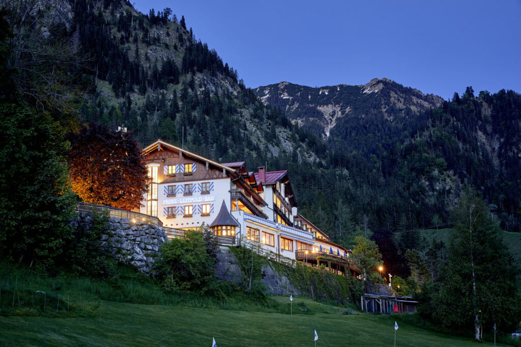 Ein großes, hell erleuchtetes Alpenhotel liegt in der Abenddämmerung an einem grasbewachsenen Hang, umgeben von hohen Bäumen und mit schneebedeckten, bewaldeten Bergen im Hintergrund. Der Himmel darüber ist klar und tiefblau.