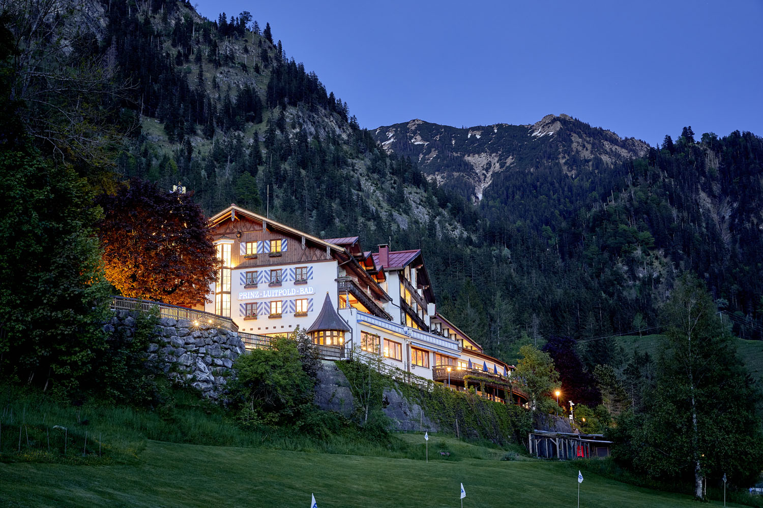 Ein großes, hell erleuchtetes Alpenhotel liegt in der Abenddämmerung an einem grasbewachsenen Hang, umgeben von hohen Bäumen und mit schneebedeckten, bewaldeten Bergen im Hintergrund. Der Himmel darüber ist klar und tiefblau.