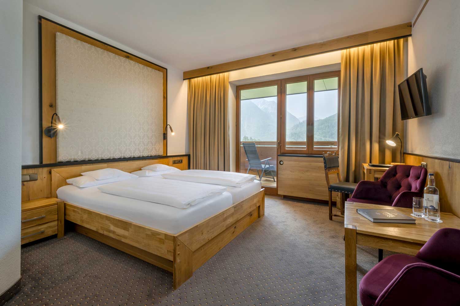 Zimmer im Modern Hotel Prinz-Luitpold-Bad mit einem Doppelbett aus Holz, Nachttischen, einem violetten Sessel, einem Schreibtisch, einem Flachbildfernseher und einem Fenster mit Blick auf die Berge. Genießen Sie die frische Luft auf dem kleinen Balkon mit Stühlen im Freien.