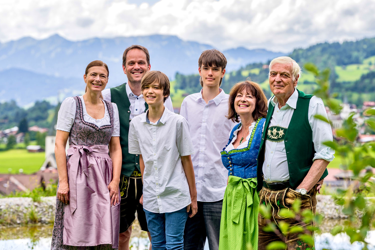 Eine sechsköpfige Familie – drei Erwachsene und drei Jugendliche – steht im Freien vor einer Bergkulisse. Einige tragen traditionelle bayerische Tracht, andere weiße Hemden; alle lächeln an diesem bewölkten Tag.