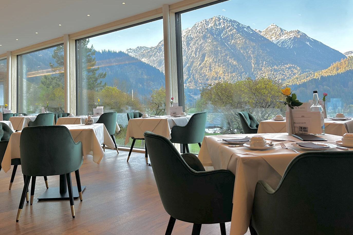Ein modernes Restaurant im Hotel Prinz-Luitpold-Bad mit grünen Stühlen und weißen Tischdecken lädt zum Essen ein. Durch die großen Fenster bietet sich ein malerischer Blick auf die Berge mit schneebedeckten Gipfeln und Bäumen im Hintergrund.