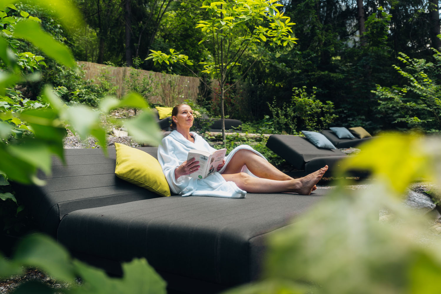 Eine Person in einem weißen Gewand entspannt sich auf einem Liegestuhl im Freien, liest ein Buch und ist von Grün und Natur umgeben. Die Szene strahlt Ruhe aus; im Hintergrund sind weitere Liegestühle zu sehen.