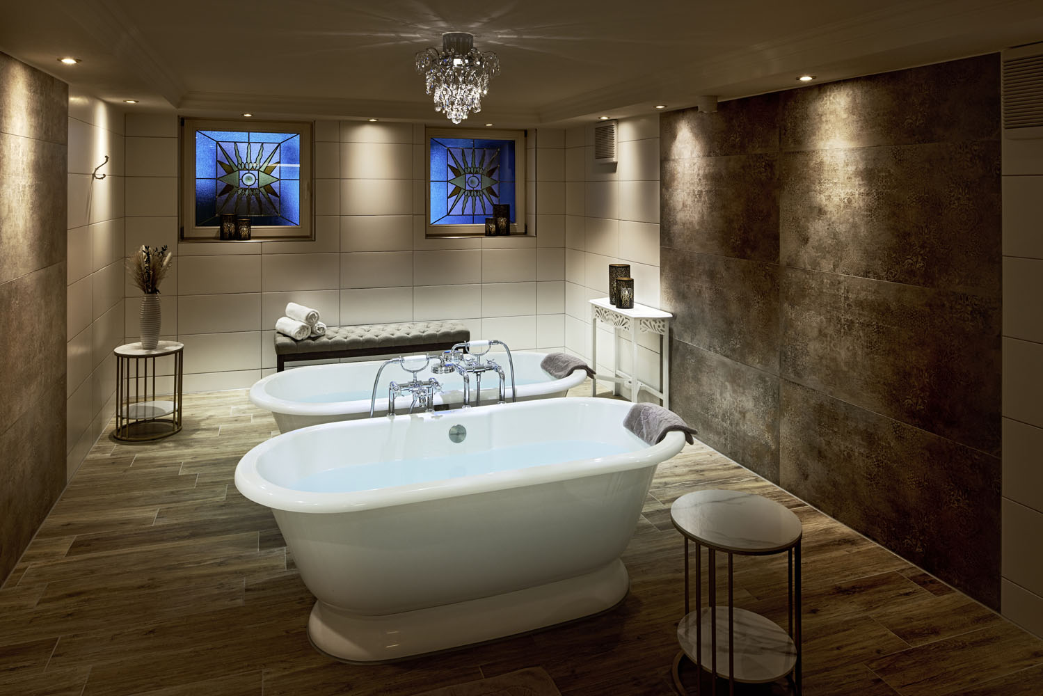 Ein luxuriöses Badezimmer mit zwei weißen freistehenden Badewannen, einem kleinen runden Beistelltisch, einer Bank unter zwei Buntglasfenstern, gefliesten Wänden und Böden, sanfter Beleuchtung und einem Kronleuchter an der Decke.