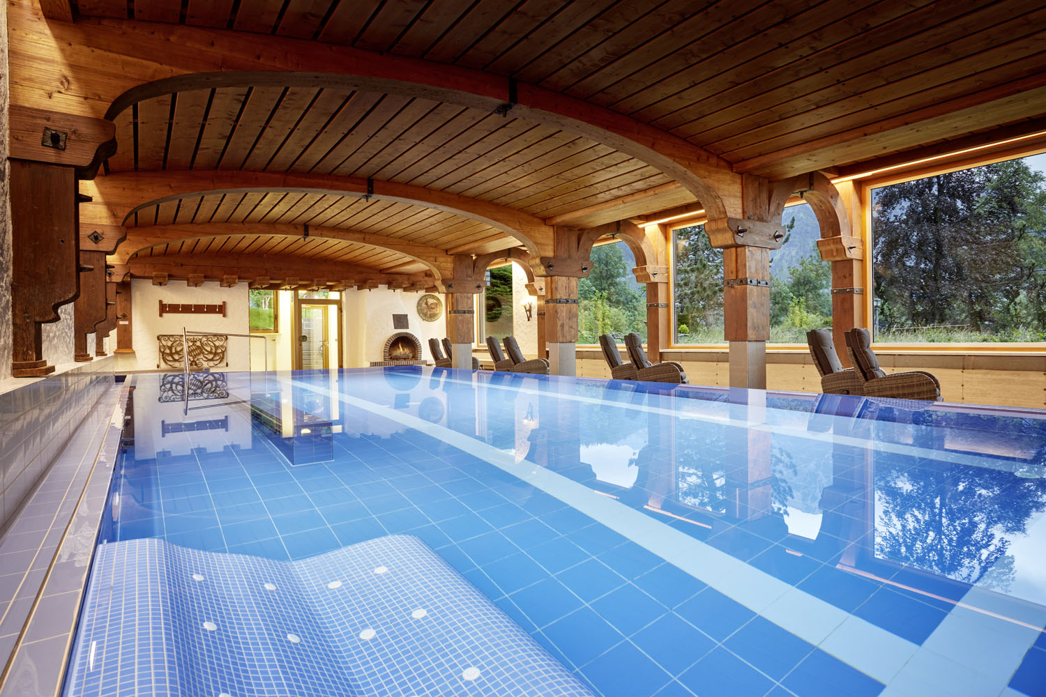 Ein Hallenbad mit blauen Fliesen, einer Holzdecke und Holzsäulen, Liegestühlen am Beckenrand, großen Fenstern mit Blick auf die grüne Umgebung draußen sowie einer warmen Stimmungsbeleuchtung, die eine entspannte Atmosphäre schafft.
