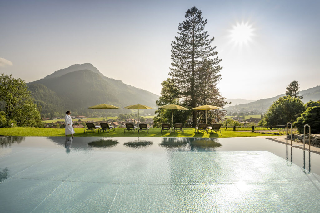 Ein ruhiger Infinity-Pool im Freien bietet bei Sonnenaufgang einen Blick auf eine malerische Berglandschaft. Liegestühle und gelbe Sonnenschirme säumen die Rasenfläche, und eine Person in einem weißen Bademantel steht am Beckenrand und genießt die Aussicht.