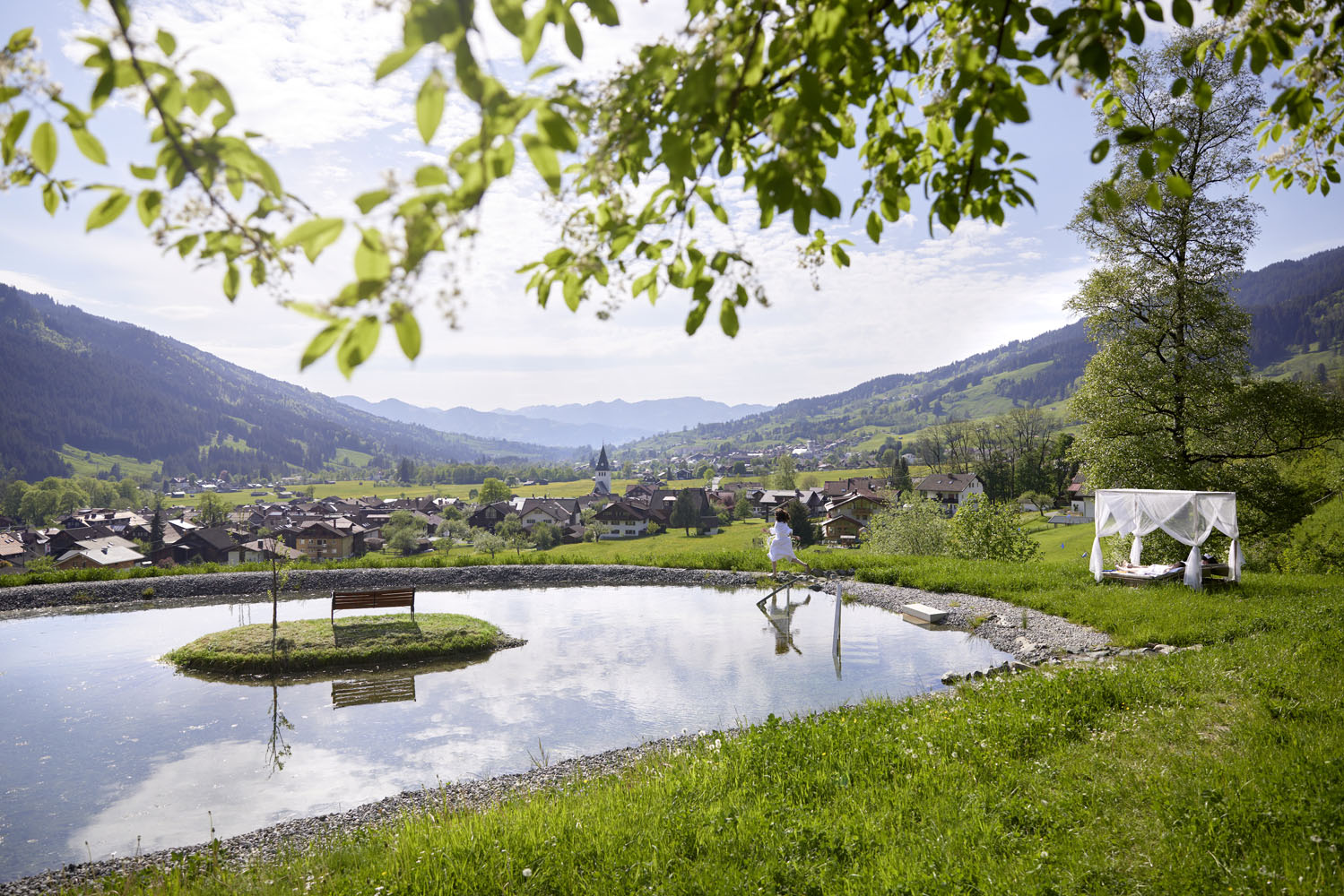 Ein malerischer Teich, in dem sich der Himmel und die umliegenden Hügel spiegeln, mit einer Bank auf einer kleinen Insel. Ein Paar steht am Teich, und auf der Wiese steht ein weißes Pavillon mit Stühlen, von dem aus man auf ein uriges Dorf im Tal blickt.