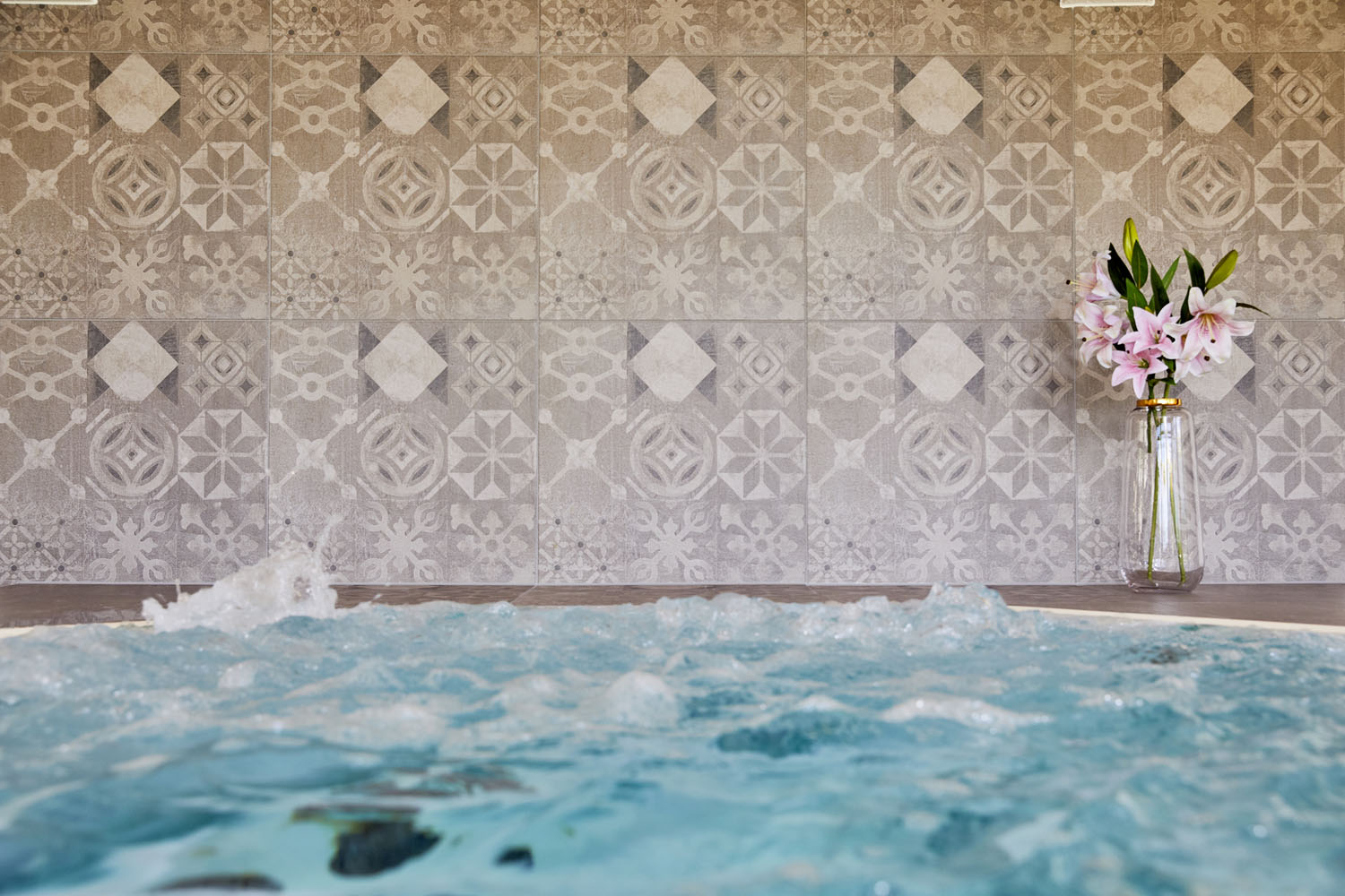 Im Vordergrund ein sprudelnder Whirlpool mit klarem, blauem Wasser, der vor einer gemusterten Fliesenwand steht. Auf der rechten Seite, nahe der Wand, steht eine hohe Glasvase mit rosa Lilien.