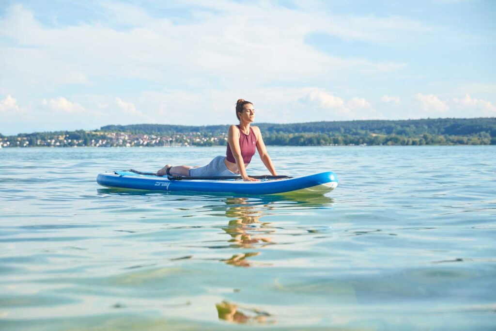 Eine Frau macht Yoga auf einem Paddleboard in ruhigem, klarem Wasser unter blauem Himmel, während im Hintergrund grüne Hügel und eine Stadt zu sehen sind.