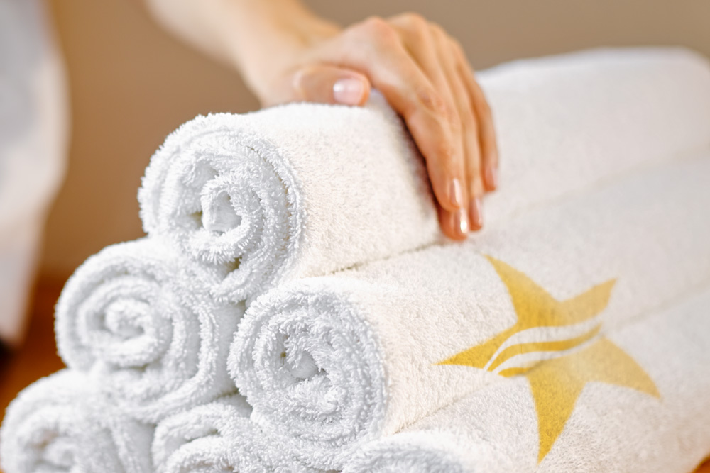 Eine Hand ordnet einen Stapel aufgerollter weißer Handtücher – eines davon mit einem goldenen Stern- und Wellenmuster – vor einem neutralen Hintergrund an – ein ruhiges Spiegelbild der Prinzipien von Wellness Stars.
