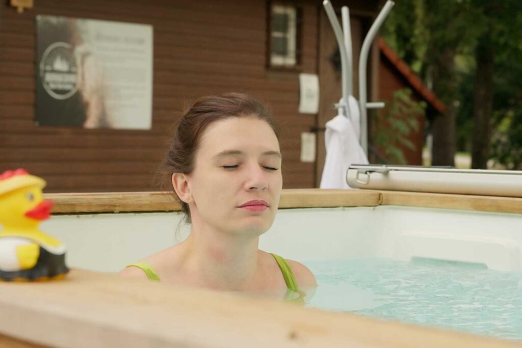 Eine Frau mit geschlossenen Augen entspannt sich in einem Whirlpool im Freien, auf dessen Rand eine Gummiente liegt und im Hintergrund ist ein Holzgebäude zu sehen.