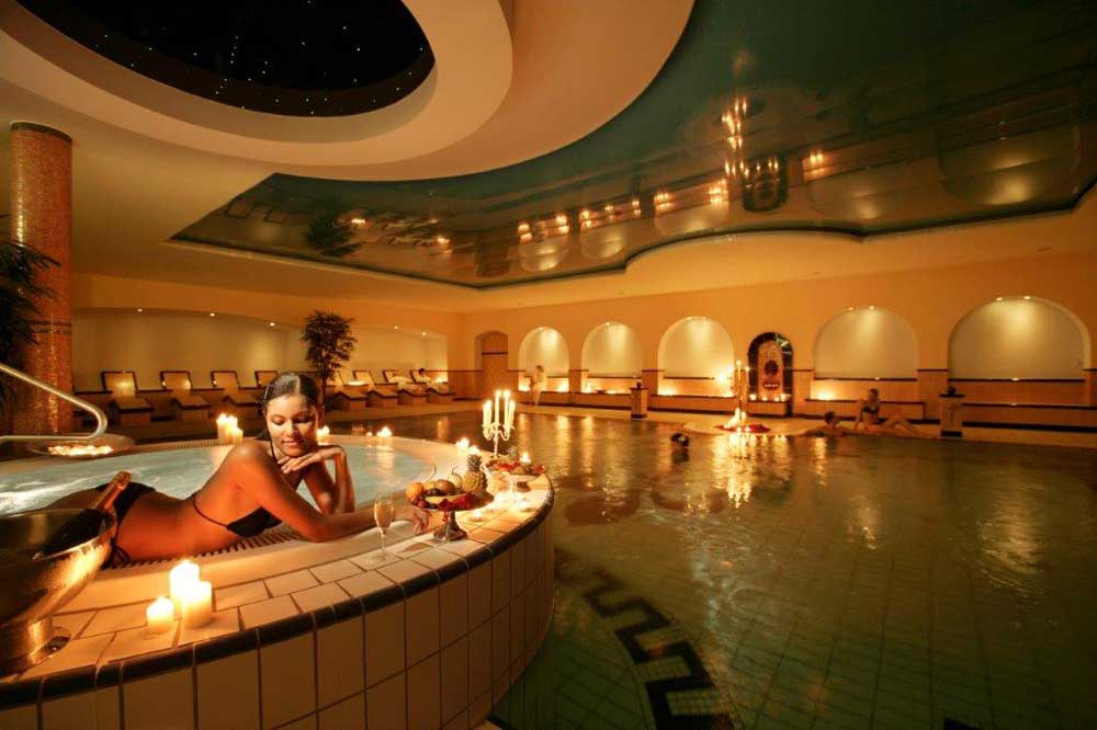 Eine Frau entspannt sich in einem von Kerzen beleuchteten Wellnessbereich, lehnt sich am Rand eines Whirlpools, neben ihr liegen Obst und Getränke, im Hintergrund sind ein großer Pool und Liegestühle zu sehen.