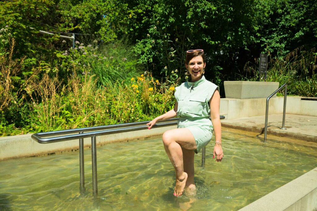 Eine Frau in hellgrüner Kleidung steht lächelnd in einem flachen Außenpool, hält sich an einem Metallgeländer fest, im Hintergrund sind Grünpflanzen und Blumen zu sehen.