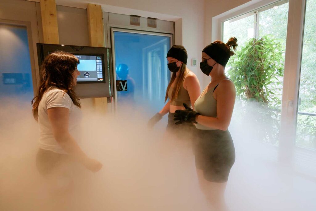 Drei Frauen, zwei davon in Sportkleidung und mit Masken, stehen in einem nebelverhangenen Raum für eine Kryotherapie. Eine Frau, die ihnen gegenübersteht, scheint Anweisungen zu geben. Sonnenlicht fällt durch große Fenster, hinter denen sich Grünflächen erstrecken.