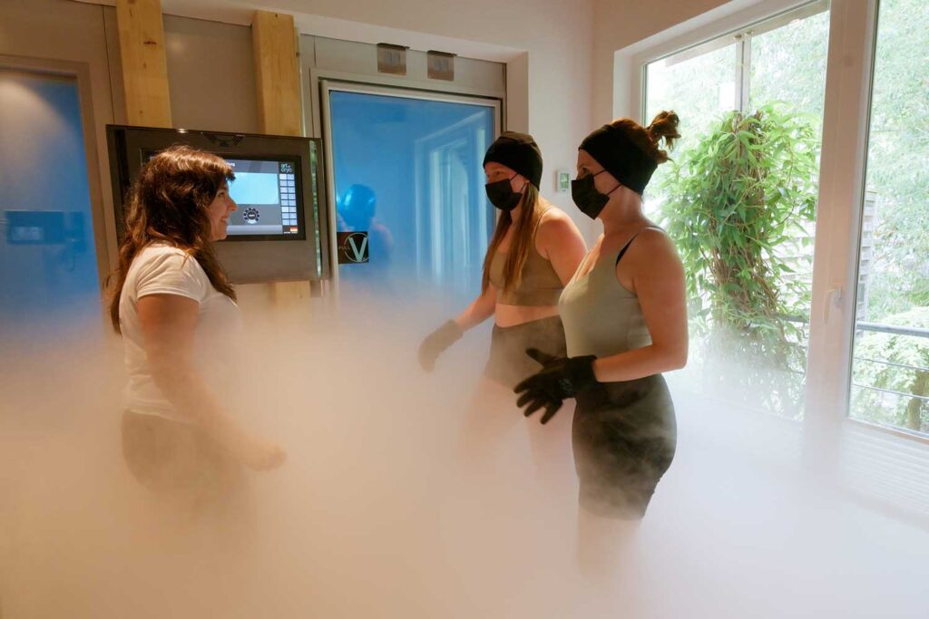Zwei Frauen in Sportkleidung, mit Mützen, Handschuhen und Masken stehen in einem nebligen Raum – möglicherweise während einer Kryotherapie-Sitzung –, während eine dritte Frau mit ihnen spricht. Sonnenlicht strömt durch große Fenster herein, hinter denen sich Grünflächen erstrecken.