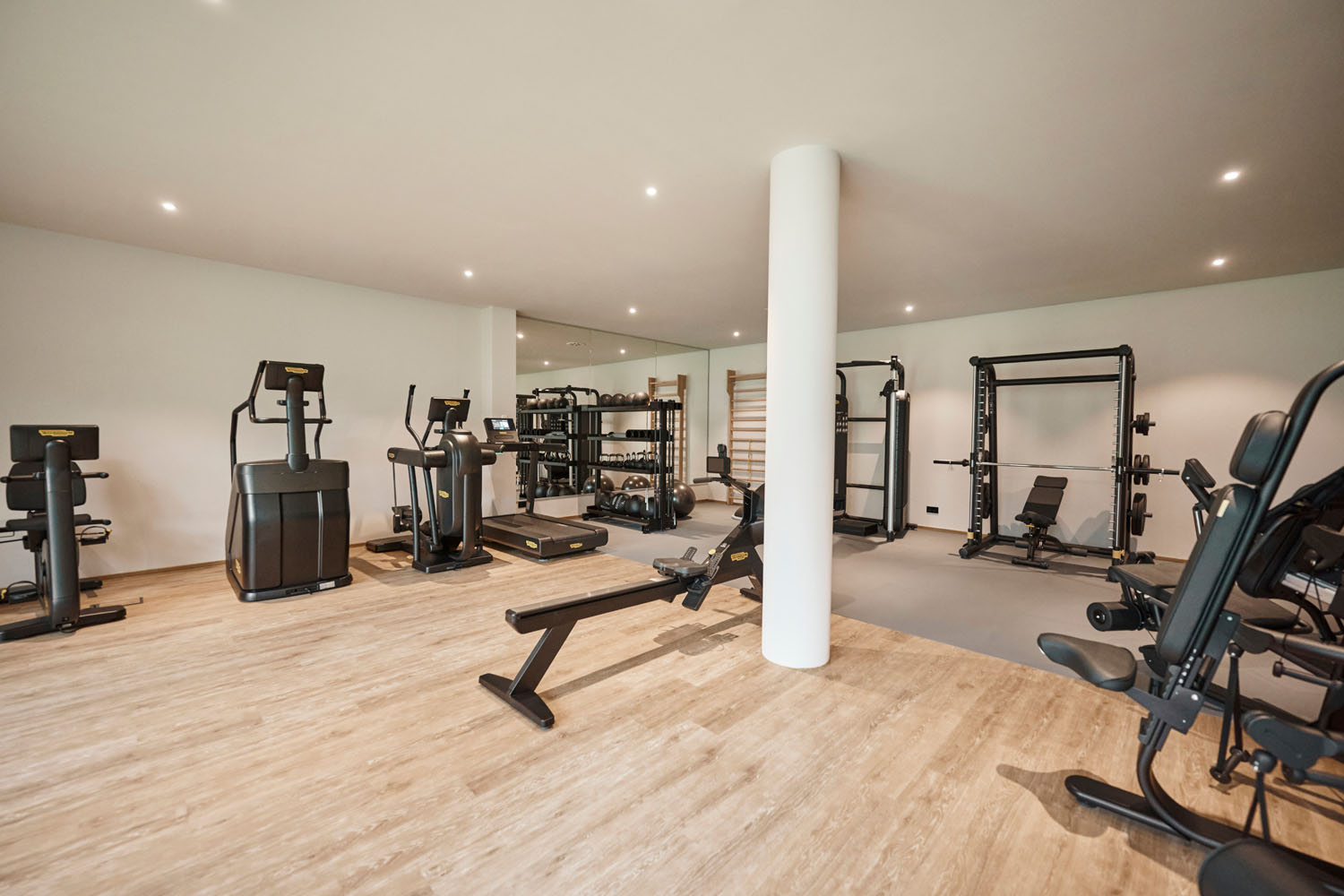 Ein modernes Fitnessstudio mit Holzboden, weißen Wänden und verschiedenen Trainingsgeräten, darunter Laufbänder, Crosstrainer, Hantelbänke, Hantelständer und Krafttrainingsgeräte, die übersichtlich um eine zentrale weiße Säule herum angeordnet sind.