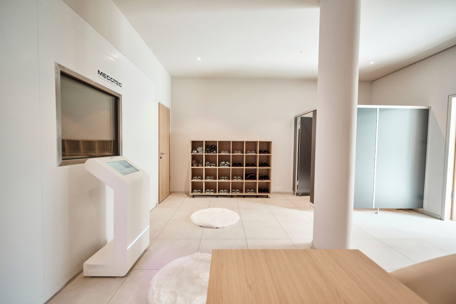 Moderne, minimalistisch gestaltete Lobby mit einem Touchscreen-Kiosk, Schuhfächern aus Holz, in denen Hausschuhe bereitliegen, runden weißen Teppichen und einer Trennwand aus Milchglas im Hintergrund. Wände und Böden sind hell und sauber.