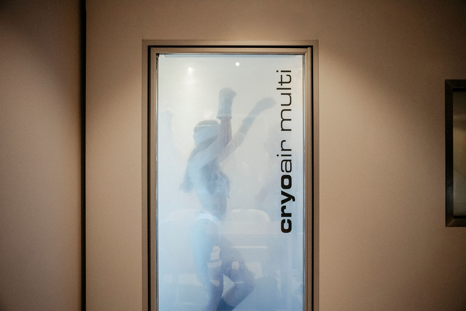 Eine Person in spärlicher Kleidung hebt in einer Kryotherapie-Kammer aus Milchglas mit der Aufschrift „cryoair multi“ die Arme.