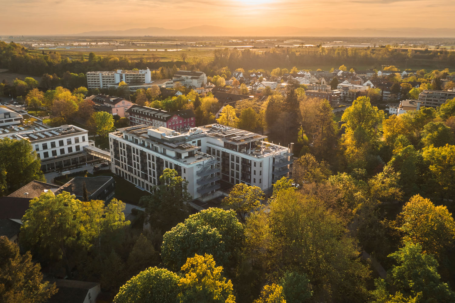 Luftaufnahme moderner, von Bäumen umgebener Gebäude, im Hintergrund eine kleine Stadt und Berge vor einem goldenen Sonnenuntergangshimmel.
