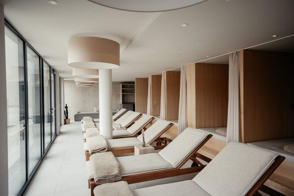 Ein moderner Wellness-Ruheraum mit mehreren gepolsterten Liegestühlen, beigen Handtüchern und Holzkabinen, dessen große Fenster für viel Tageslicht sorgen.